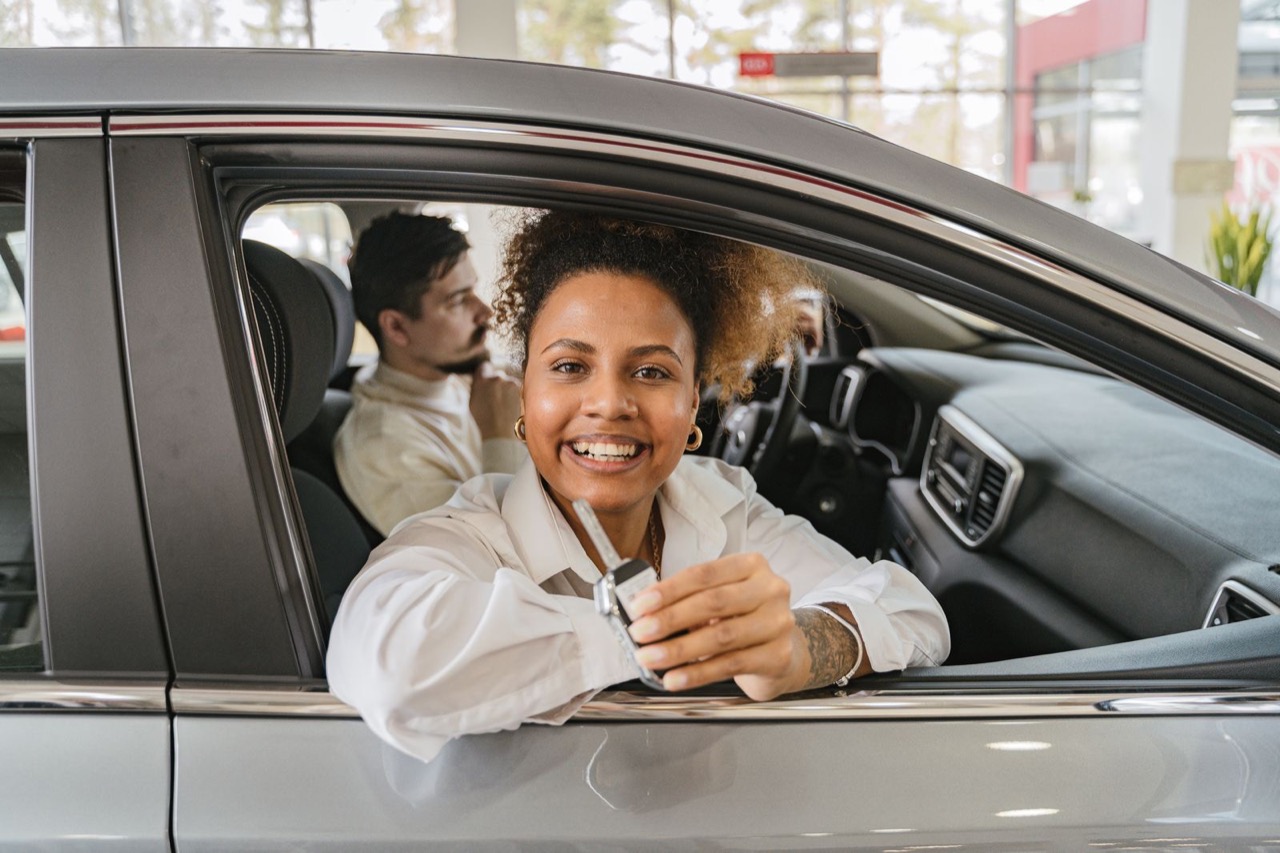 A smiling woman leaning out of a car window holding up a key, with a salesperson in the passenger seat