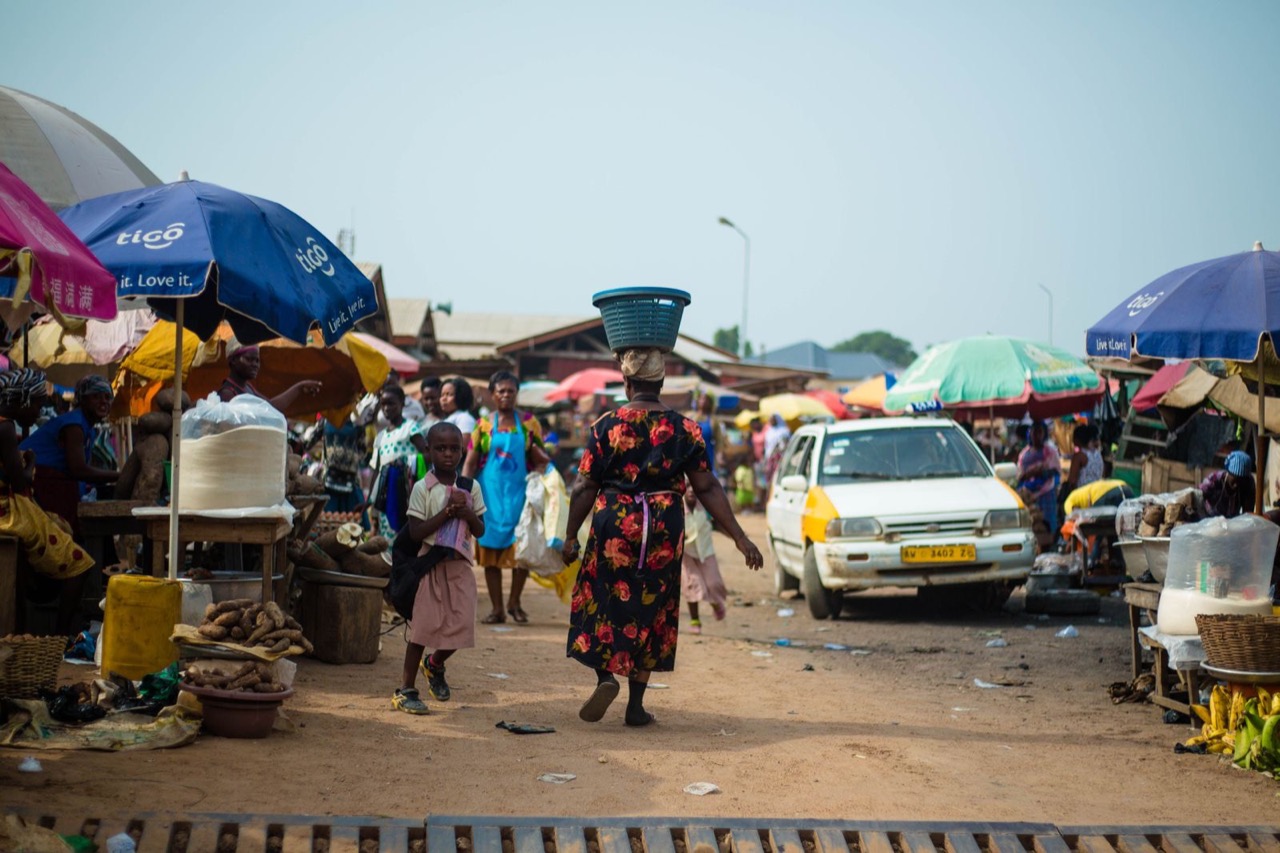 A busy Accra market with traders under coloured umbrellas, a woman carrying a basin on her head and a taxi easing through the crowd