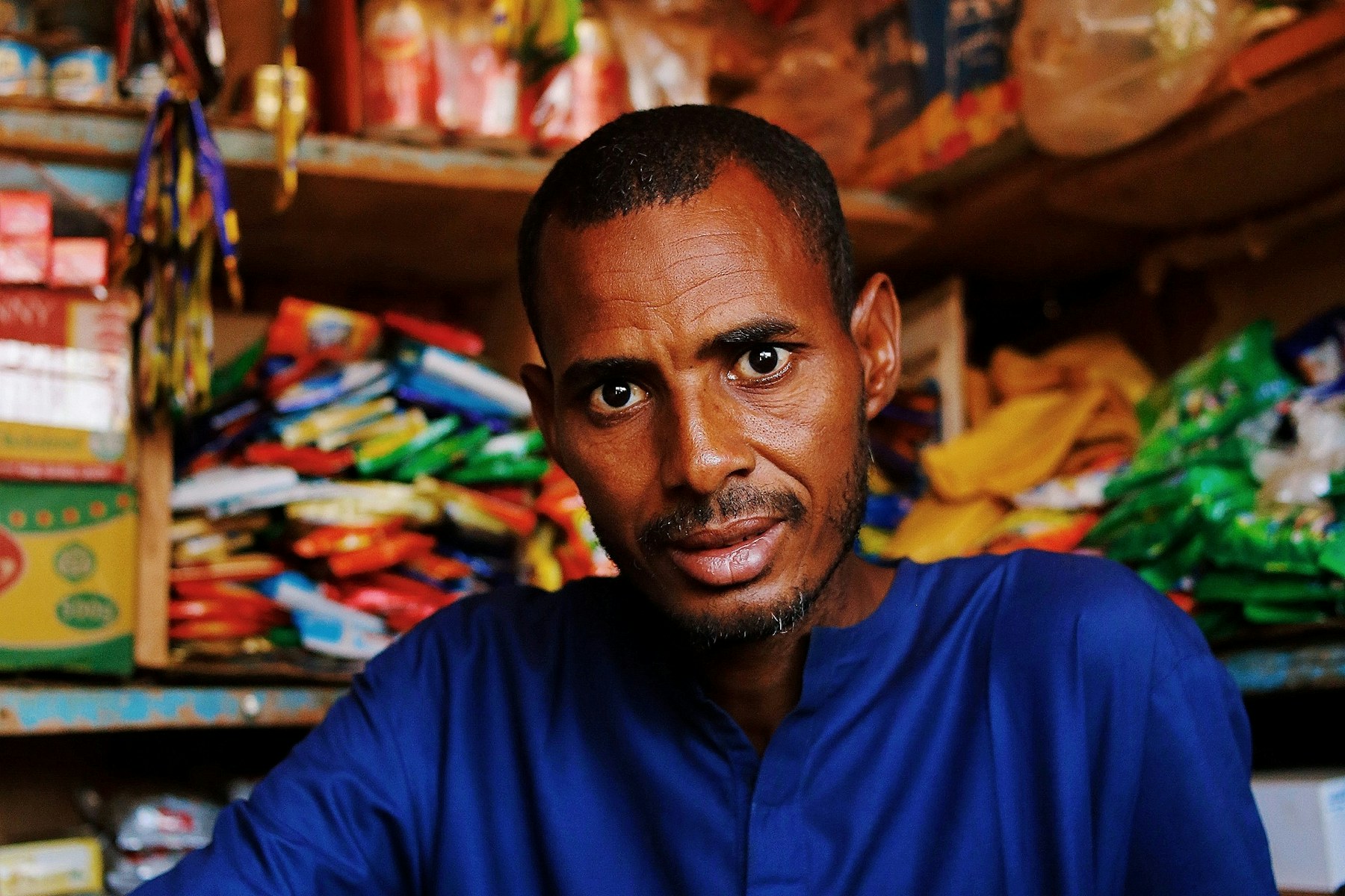 A shop owner in a blue shirt standing among the stock in his small shop