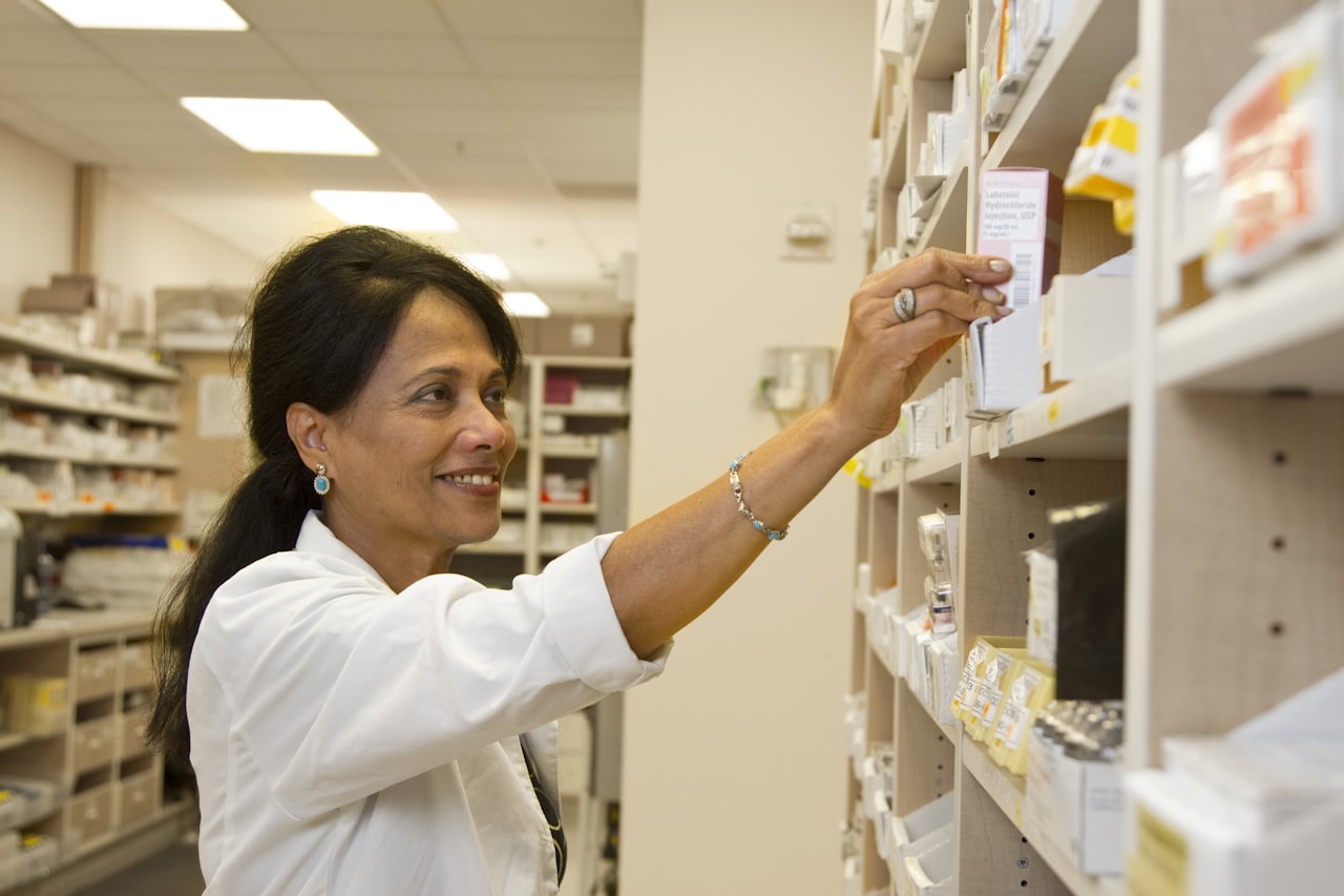 A pharmacist in a white coat reaching for a boxed medicine on a dispensary shelf